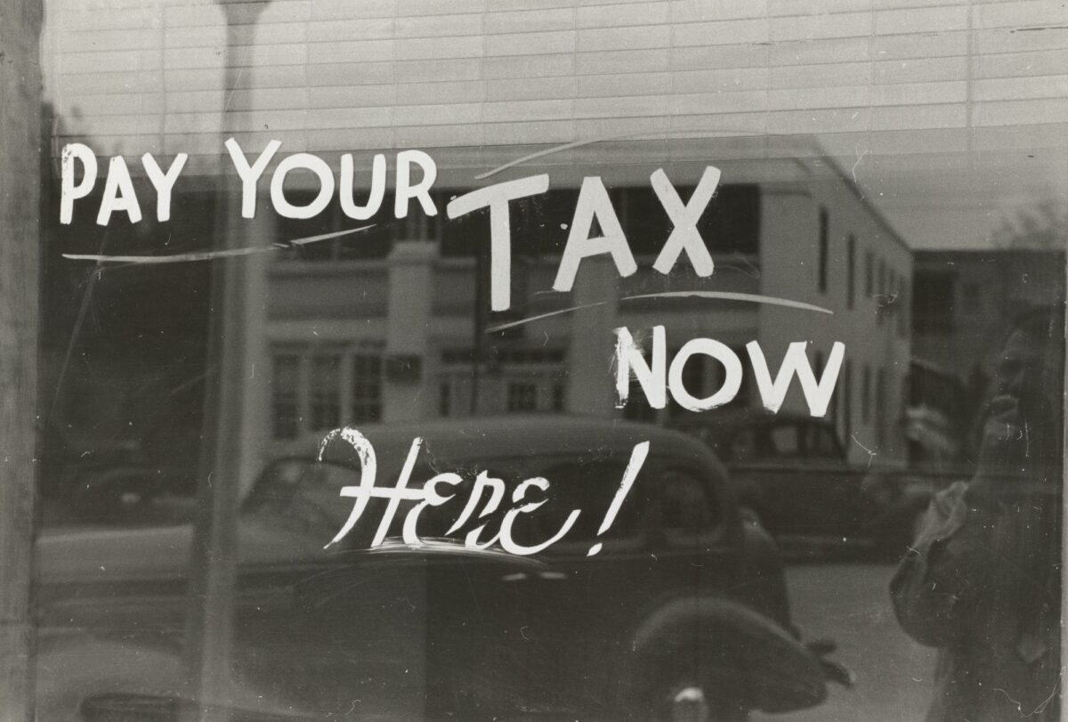 Image is vintage black and white, of a window with the words Pay Your Tax Now Here painted on the glass. IN the reflection is a building and a vintage car