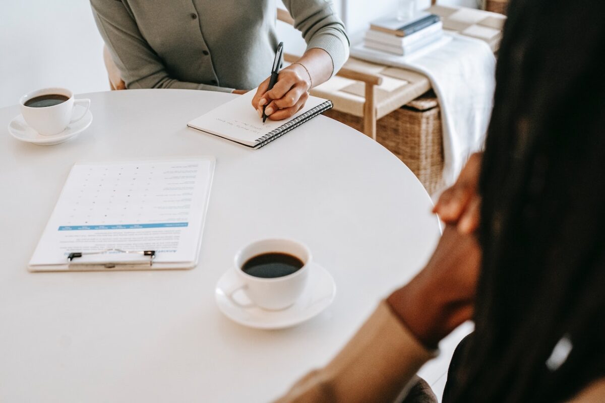 Two people sit at a plain, circular desk. One has their hands crossed, and the other is writing on a notepad. There are black coffees on the table and an interview checklist.